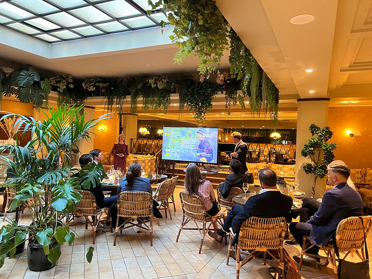 Speaker presenting to seated guests in a plant-filled restaurant hall at Quantfolio's Wealth Meetup at Sommerro, Oslo